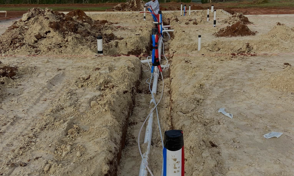 A construction site showing a trench in sandy soil with installed plumbing pipes and vertical capped conduit pipes.