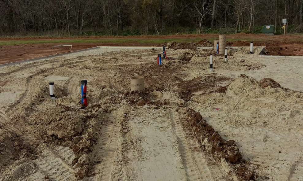 Construction site with exposed plumbing pipes protruding from a sandy, dirt-covered foundation area.