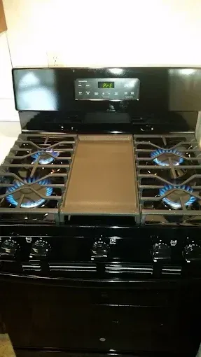 A black gas stove with four lit burners and a central griddle, featuring a digital control panel on the back panel.