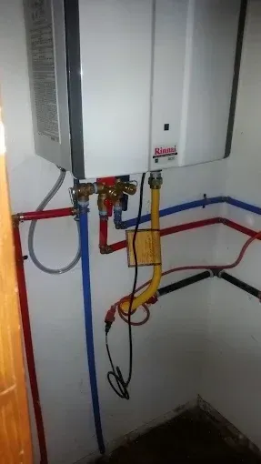 A Rinnai tankless water heater mounted on a wall with connected red, blue, and yellow gas piping and electrical wiring.