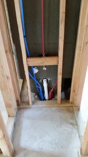 Plumbing pipes and a white drain pipe installed between wooden wall studs in a room under construction.