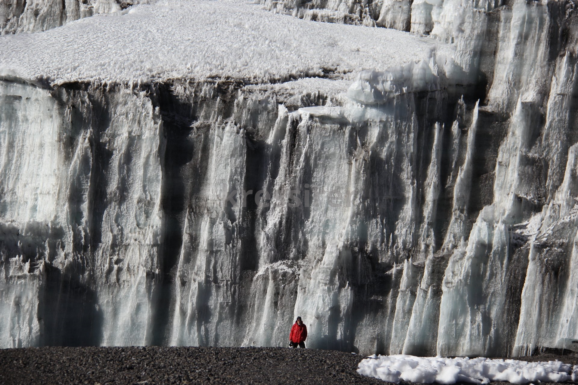 A man in a red jacket stands in front of a large wall of ice