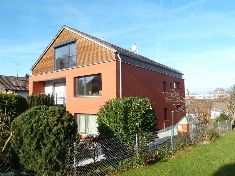 A large red house with a wooden roof is surrounded by bushes and a chain link fence.