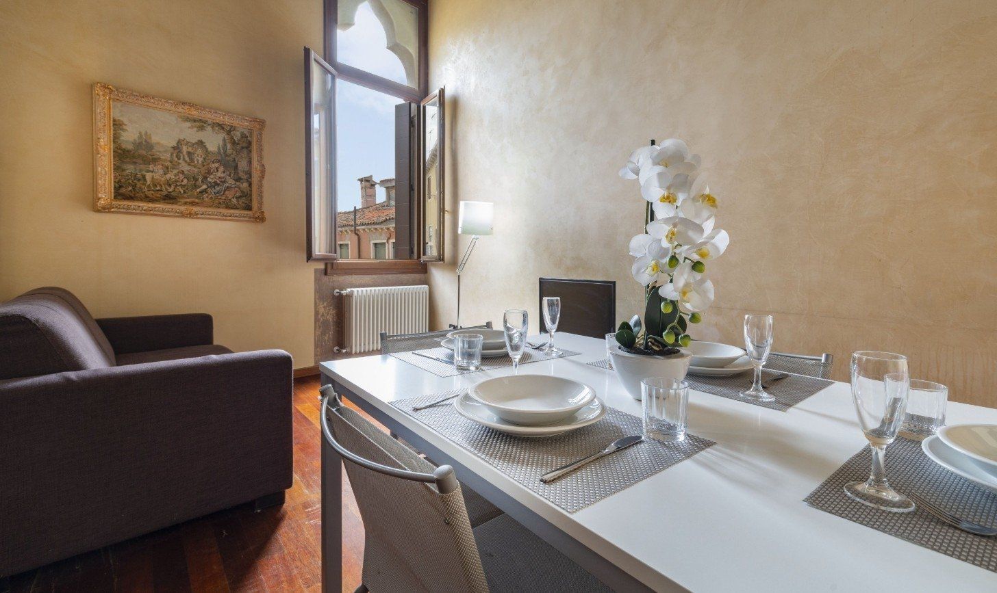 Elegant dining room with set table, white flowers, and a sunlit window beside a dark sofa.