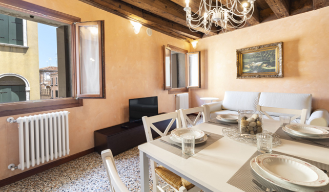 Bright dining room with white table setting, rustic wood beams, chandelier, and large window.