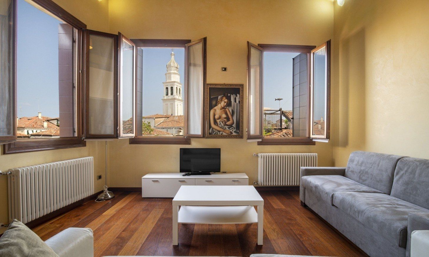 Bright living room with open windows, gray sofa, coffee table, TV, and a city tower view outside.