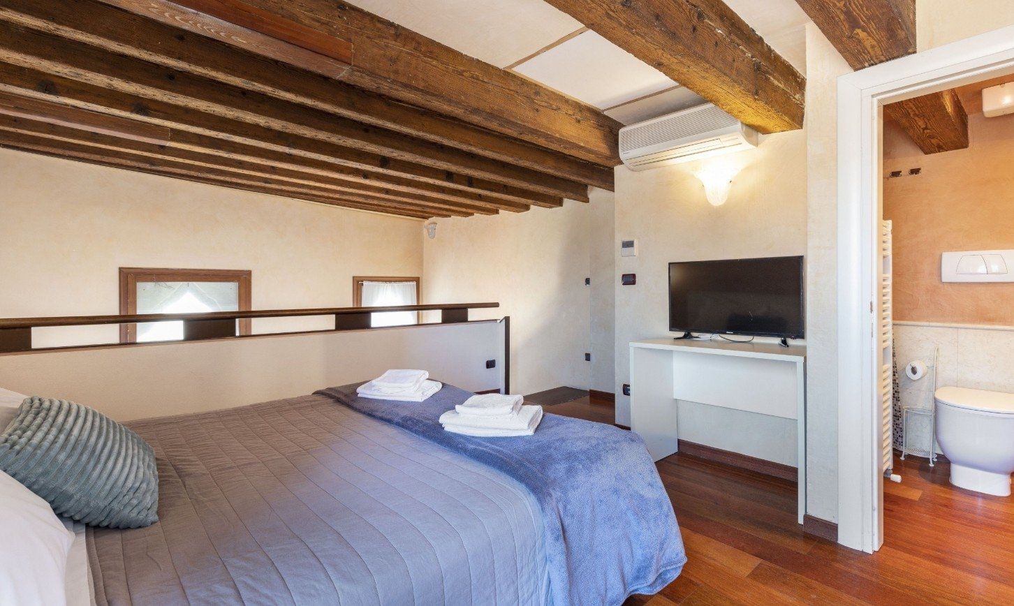 Rustic bedroom with exposed beams, gray bedspread, and adjoining bathroom visible through open doorway