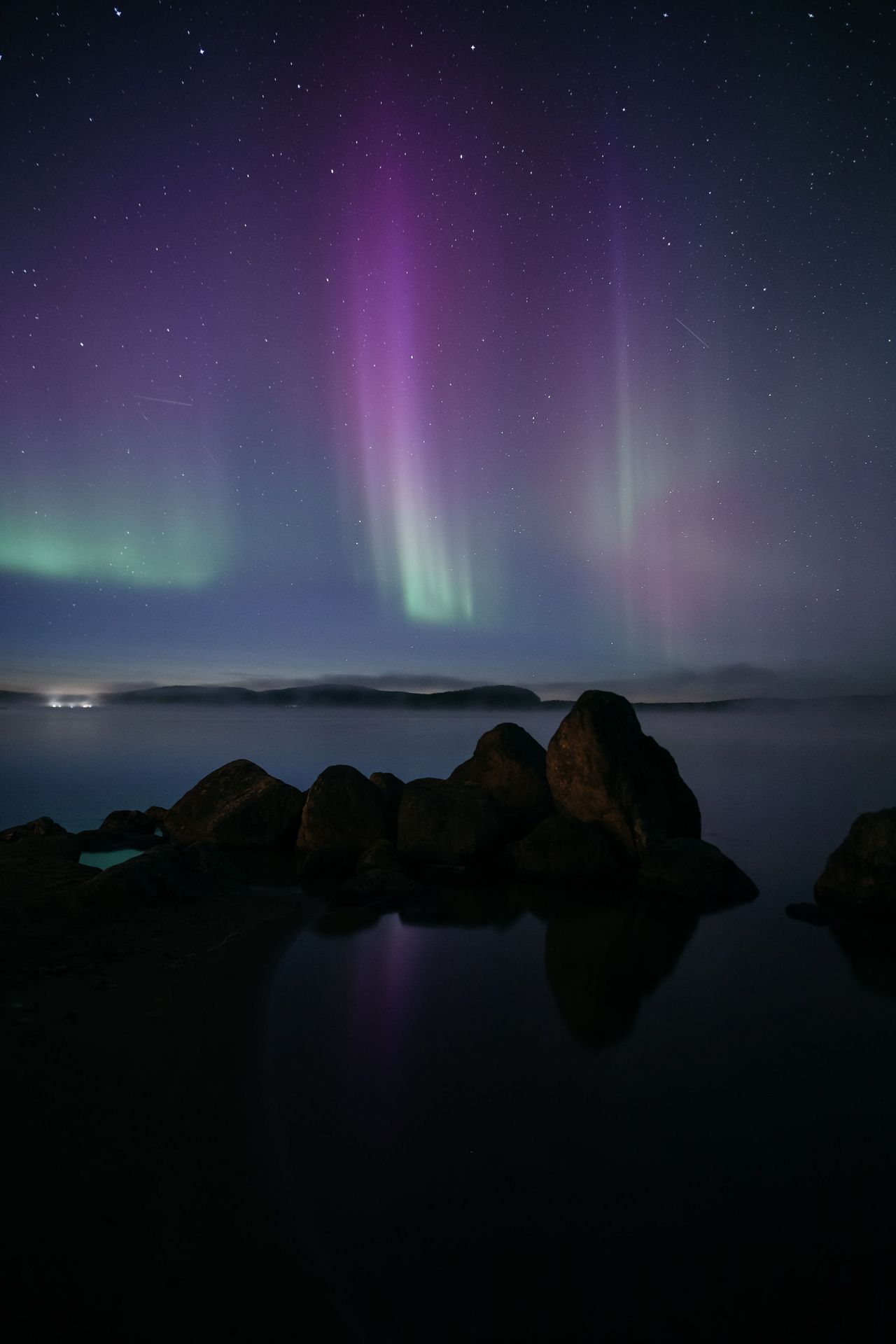 Purple and green aurora borealis lights over dark water, rocks in foreground. Starry night sky.