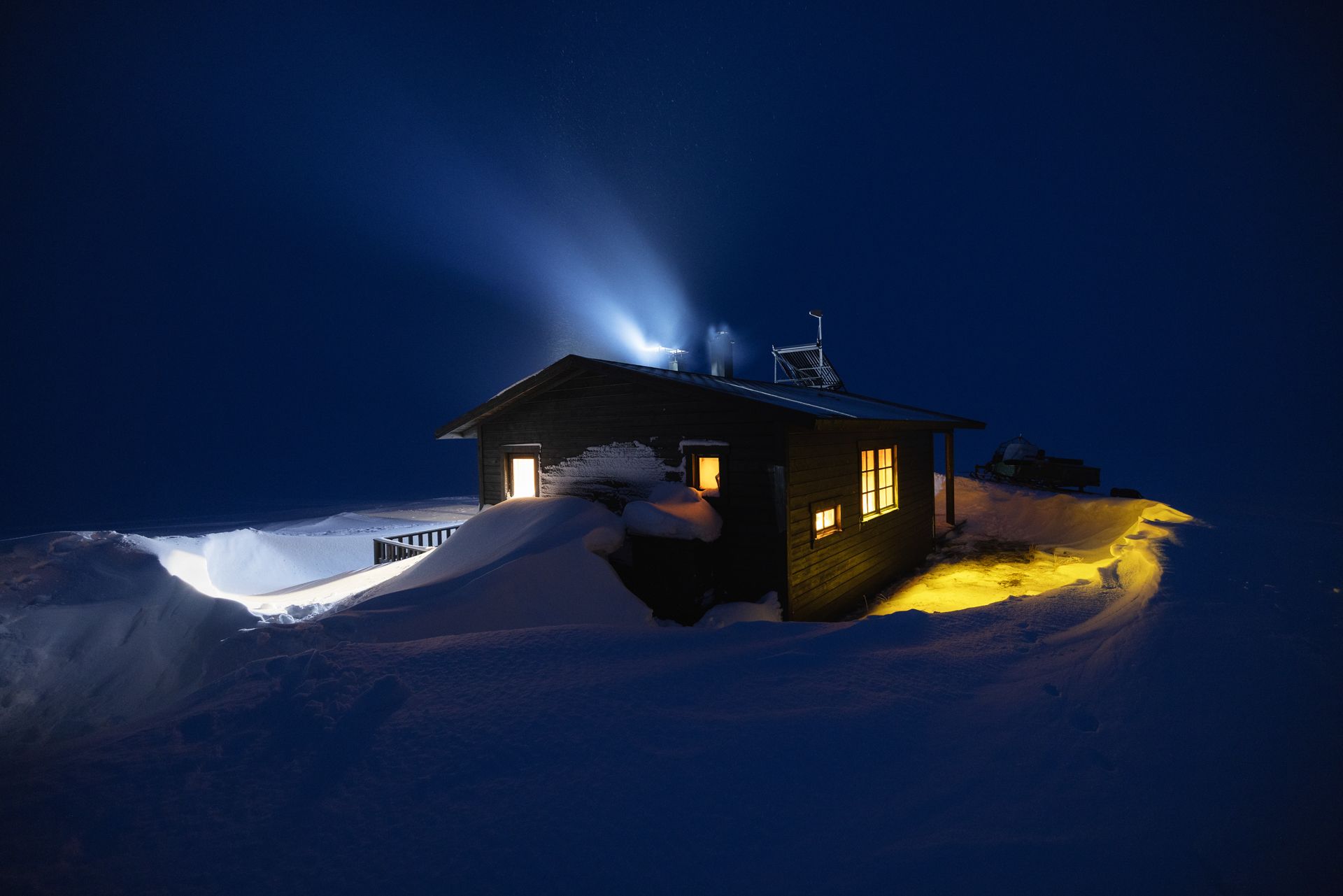 Cozy cabin in snowy landscape at night, illuminated by interior lights and a spotlight.