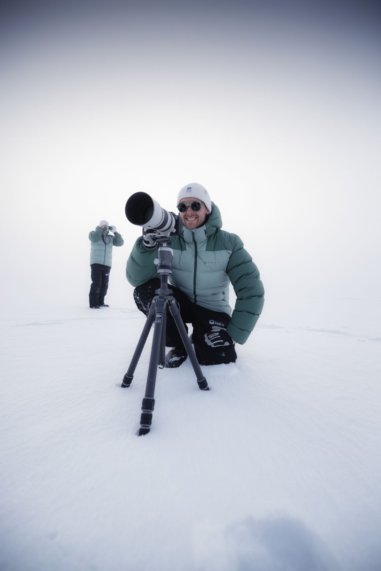 Man kneels in snow, looking through a long camera lens on a tripod. Another person is visible in the background. Snowy setting.