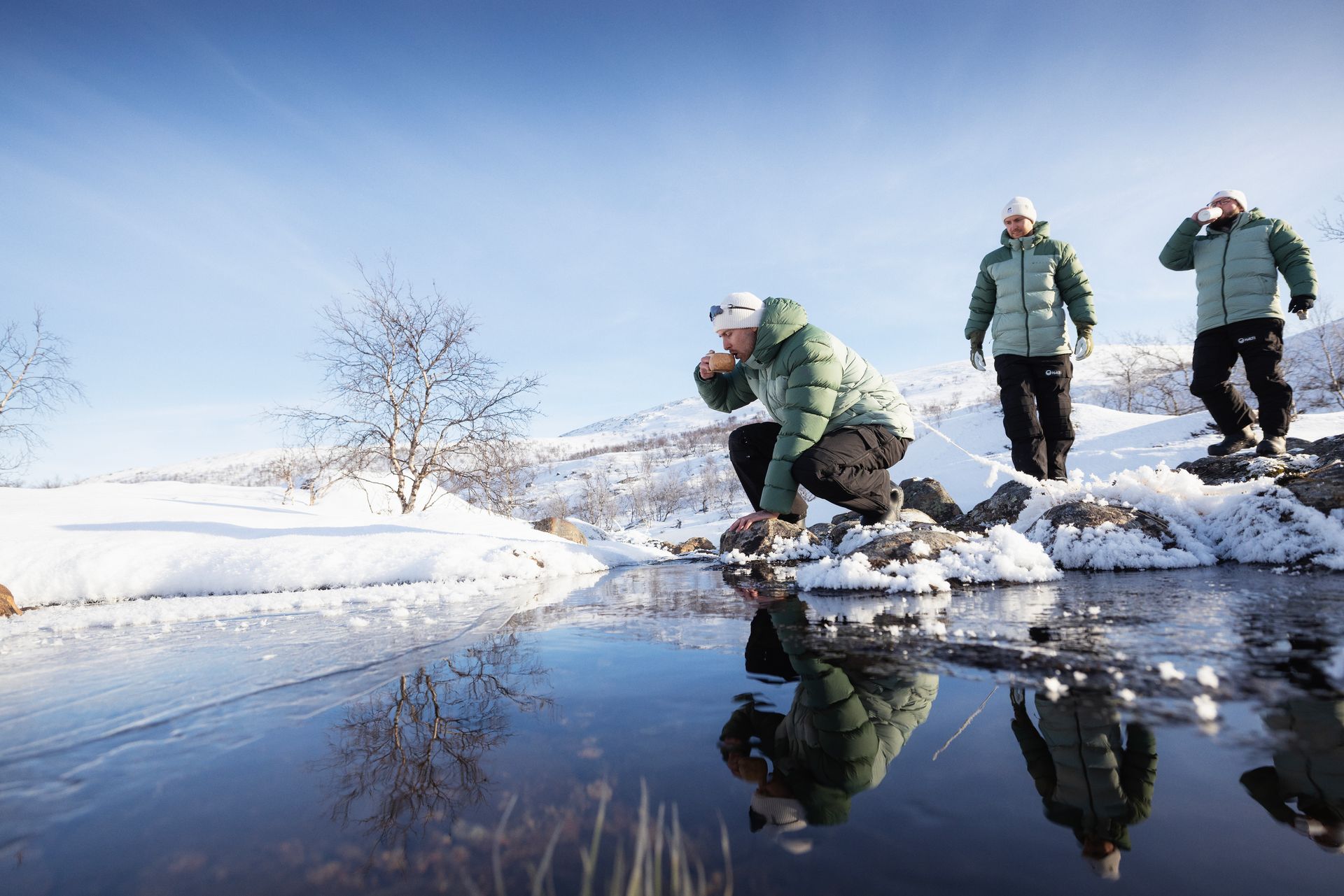 Three people by a partly frozen lake, one drinking water. Snowy landscape, blue sky.