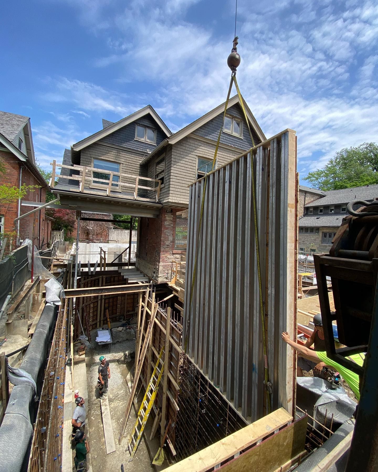 A crane lowers a large, vertical wall panel into a narrow construction trench next to a house under renovation.