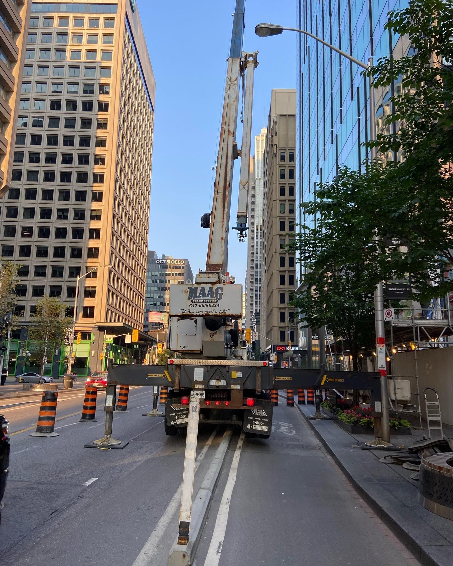A white construction crane with extended outriggers blocks a city street between two tall office buildings at sunset.