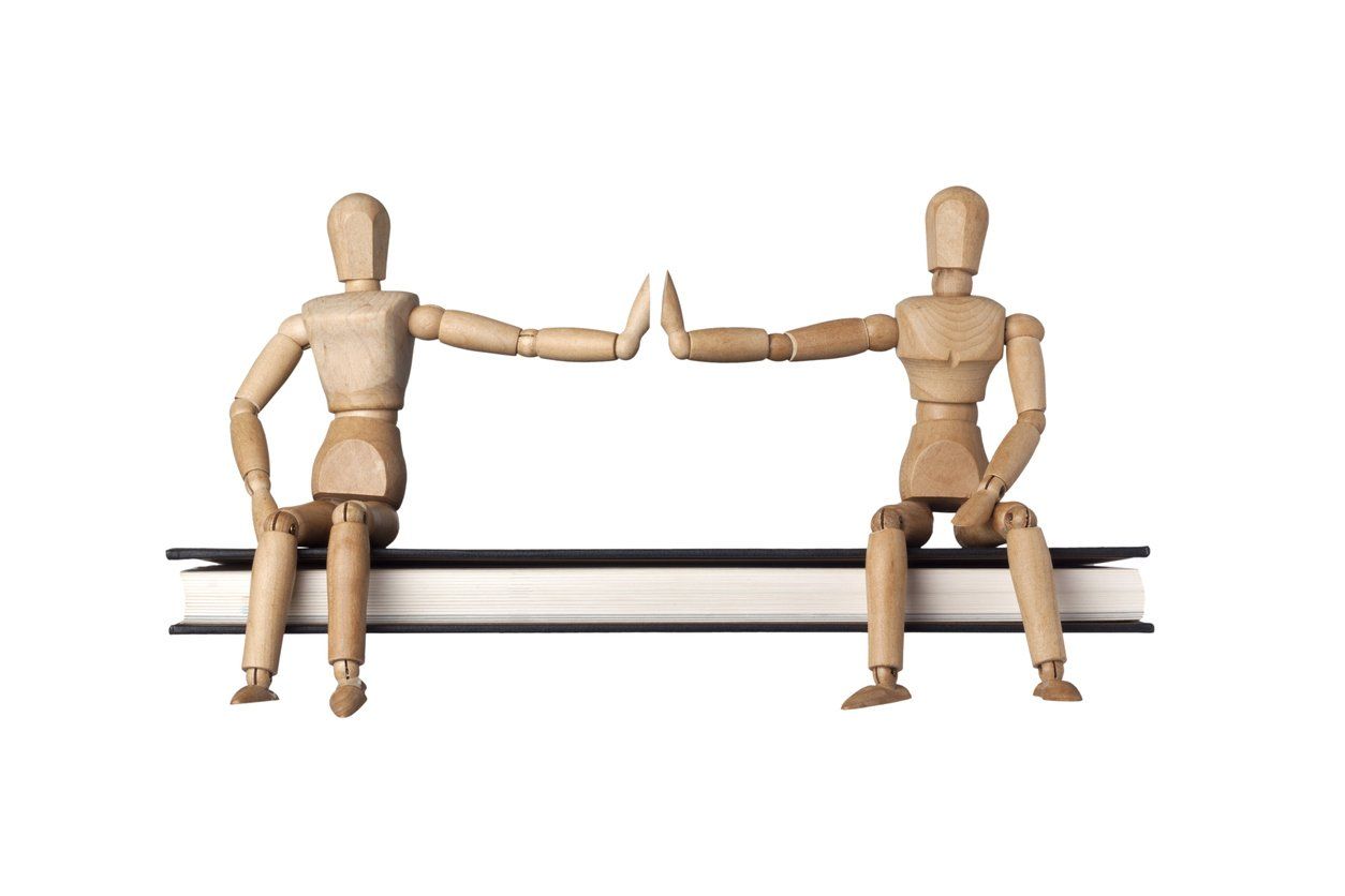 Two wooden mannequins are sitting on a book and giving each other a high five