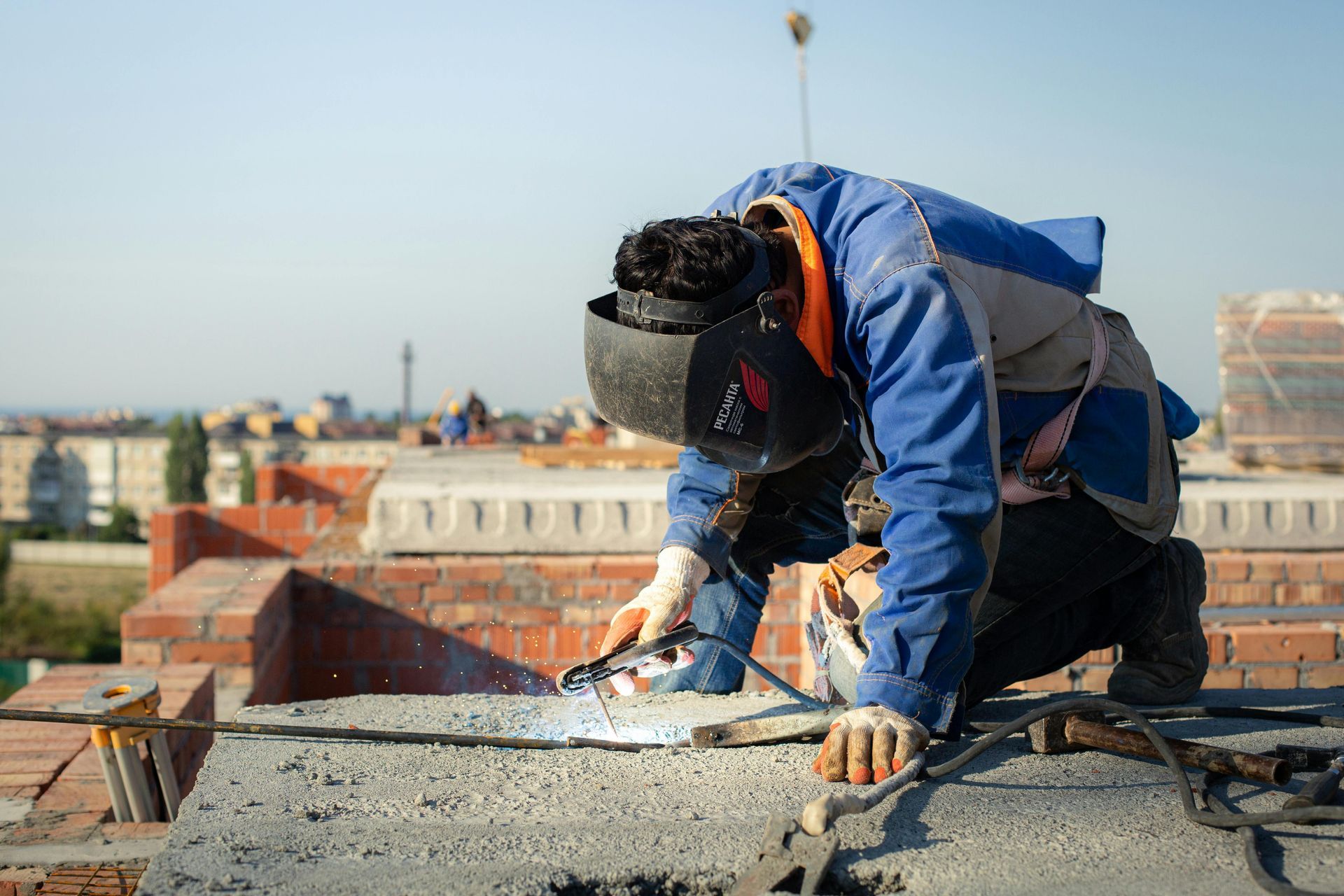 Welder wearing protective gear, welding on a rooftop with city in the background.