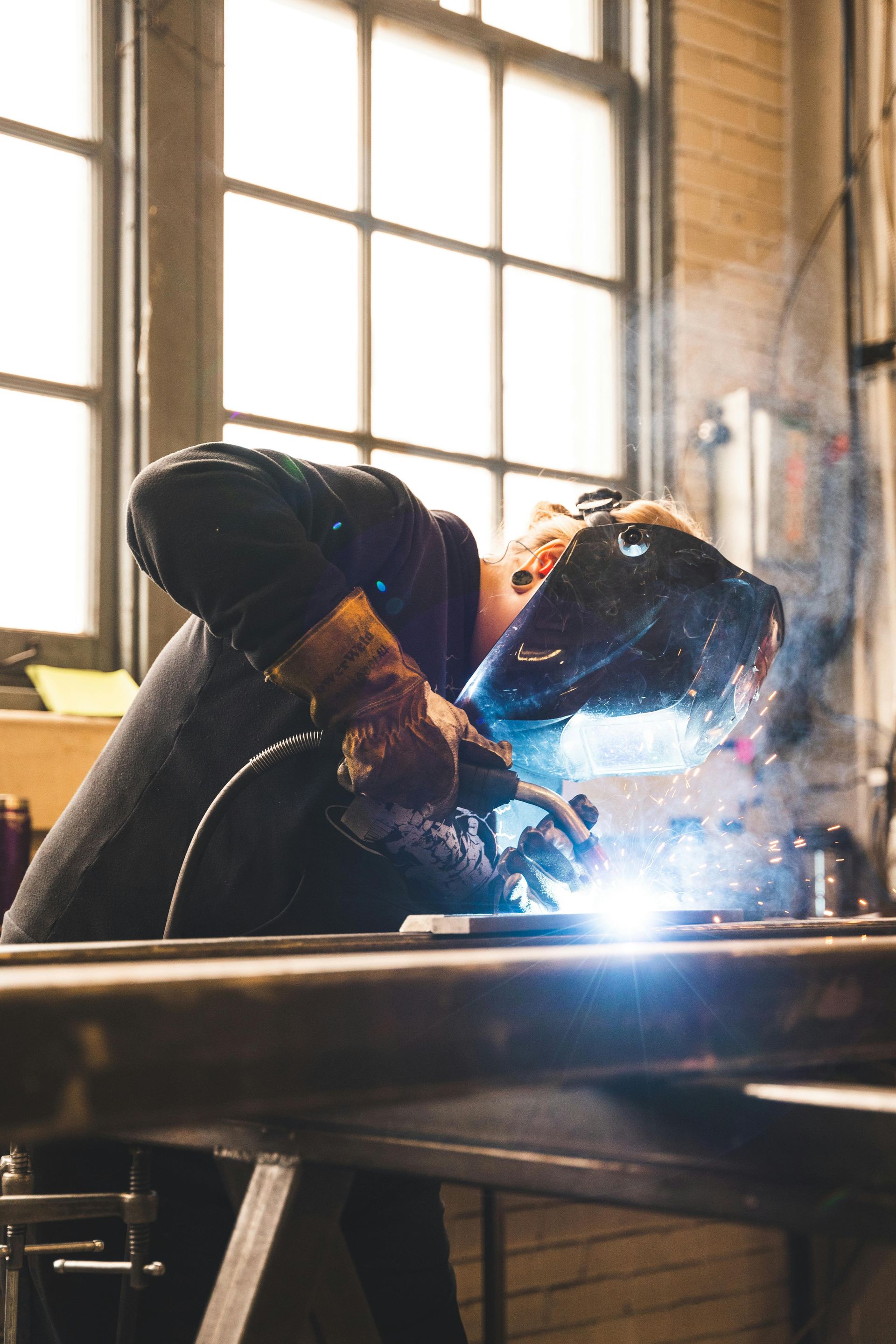 Welder wearing a helmet and gloves, working on metal in a workshop. Bright sparks illuminate the scene.