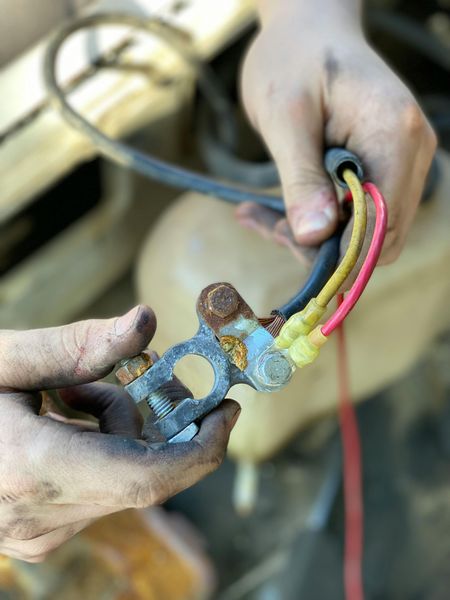 Hands holding a corroded car battery terminal with red, yellow, and black wires.