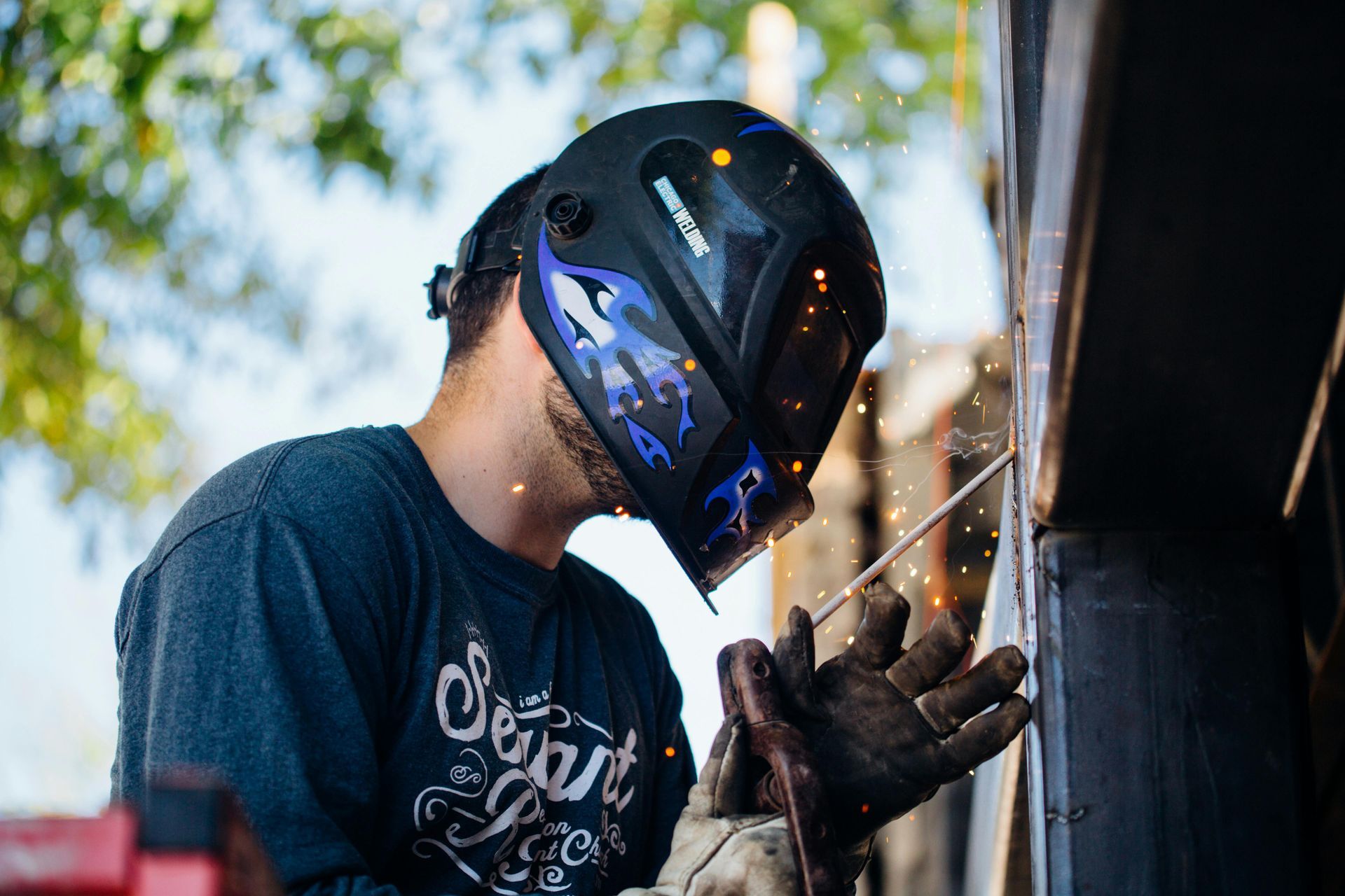 Welder wearing a dark helmet with flames, sparks flying, outdoors.