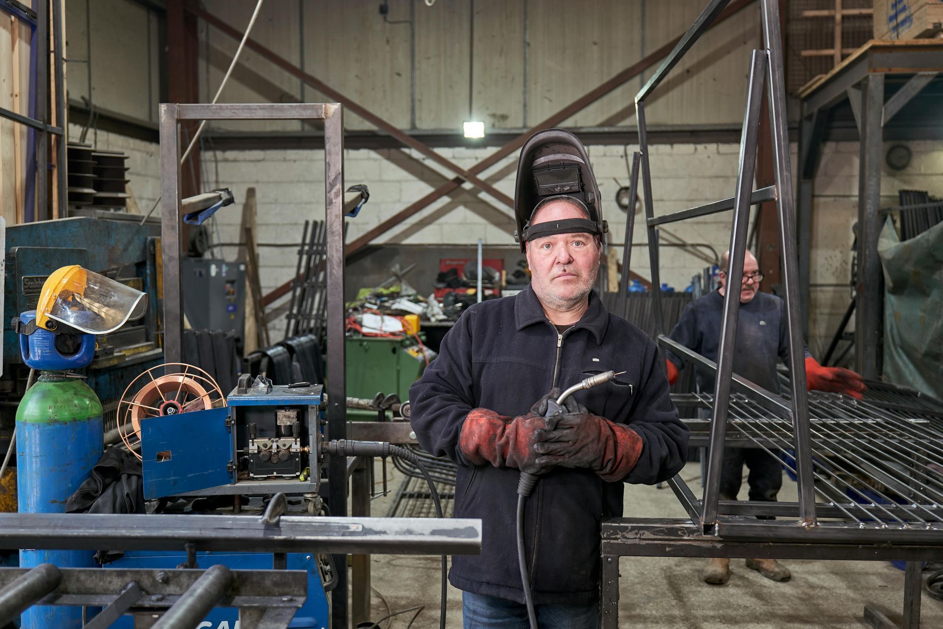 Welder in shop, holding torch, wearing helmet and gloves, standing in front of metal frame, another person in the background.