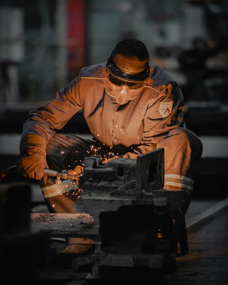 A worker wearing protective gear welds metal parts in a workshop, illuminated by bright orange sparks.