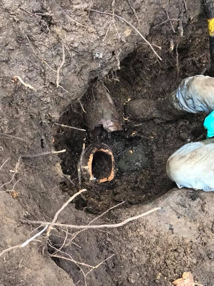 Hole dug in soil reveals a dark, rusty metal pipe surrounded by dirt and roots. Hands in gloves reach in.