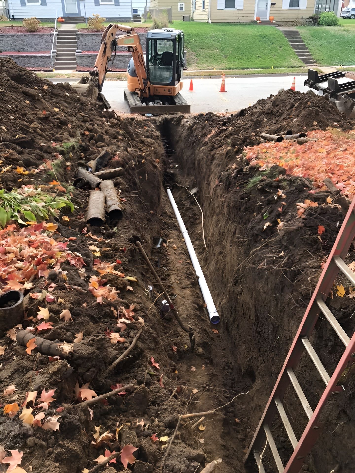 Construction site: excavator digging trench for white pipe, ladder leans on the side, residential street in the background.