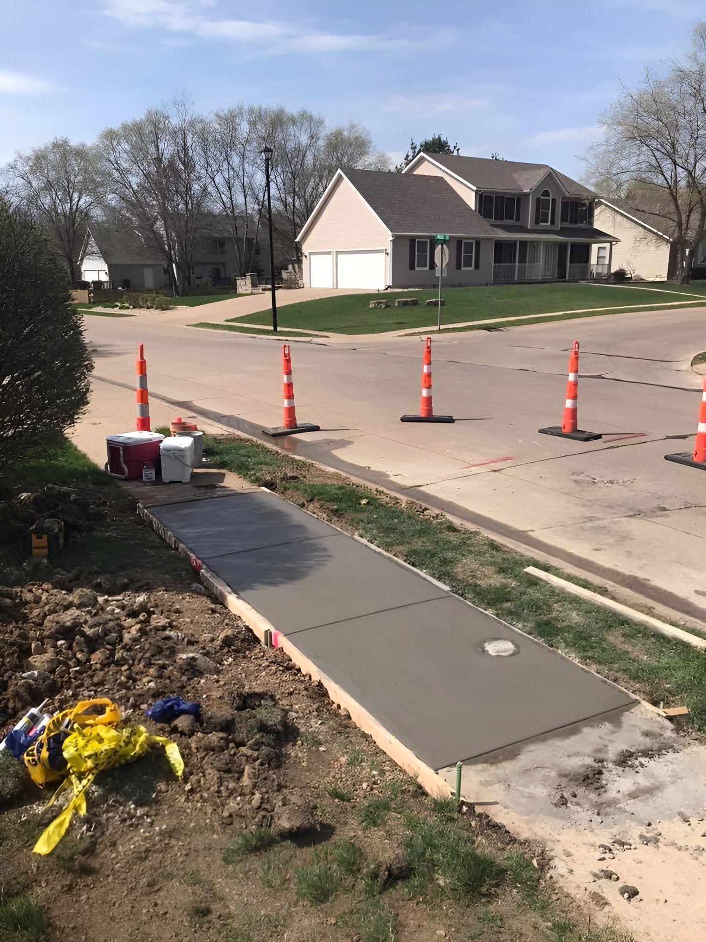 New concrete sidewalk being installed in a residential neighborhood, orange traffic cones mark the street.
