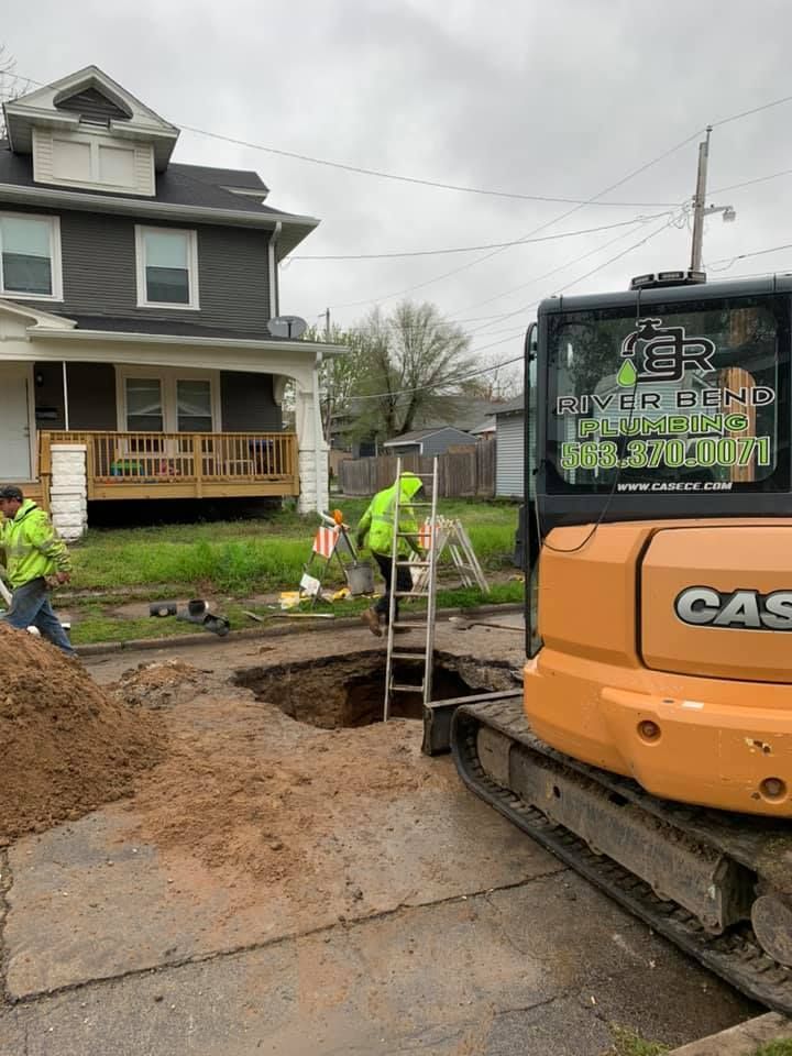 Construction workers near a hole in a street with an excavator. One worker descends a ladder. Gray house in the background.
