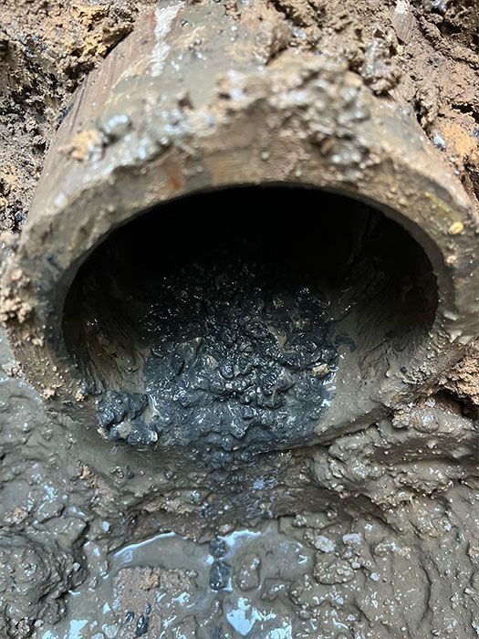 Open end of a corroded pipe filled with sludge and surrounded by wet soil.