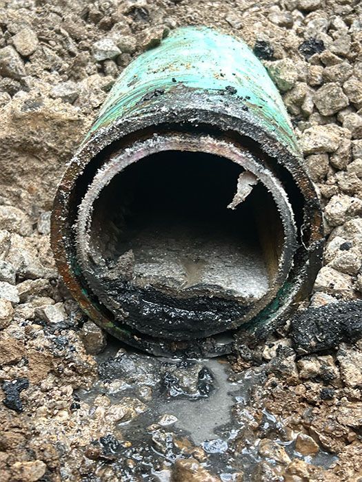 Cut-open green pipe surrounded by gravel, revealing a corroded interior and pooled liquid.