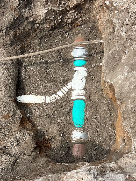 Underground pipe with a valve, painted white and wrapped in teal insulation, in a dirt trench.