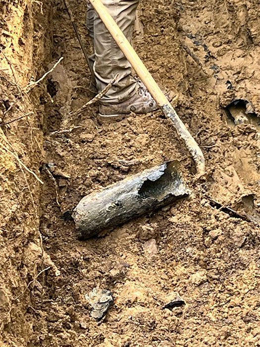 Person digging in dirt with a shovel, uncovering a cylindrical, corroded metal object.