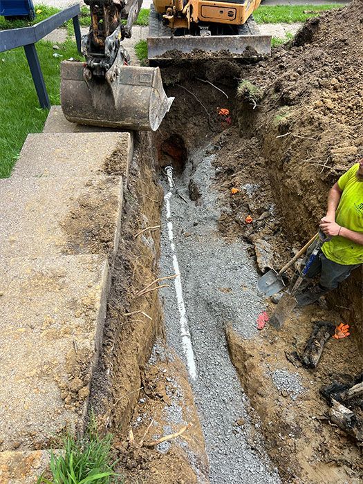 Trench dug beside a concrete structure, with a white pipe laid in gravel; backhoe and worker present.