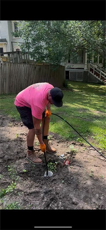 A person in a pink shirt works with a tool in the dirt of a yard, near a fence and a house.