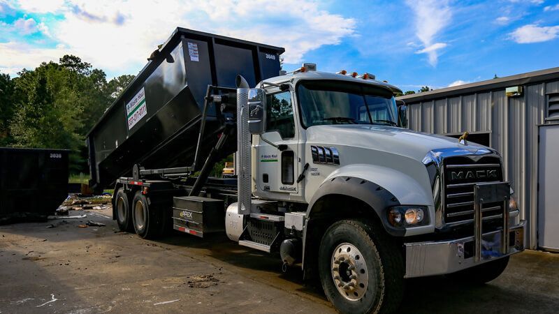 White Mack dump truck with raised black bed parked in a yard near a building, side view