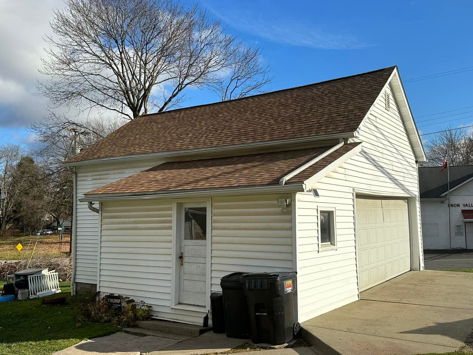A white garage with a brown roof and a trash can in front of it.