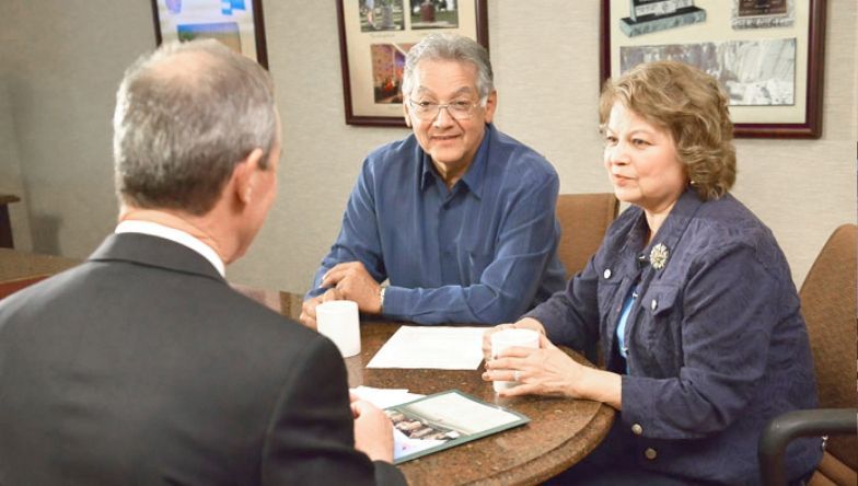 A man and a woman are sitting at a table talking to a man in a suit.