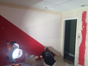 Room with red accent wall, exposed wall patches, and a person working near a lamp and tools on the floor