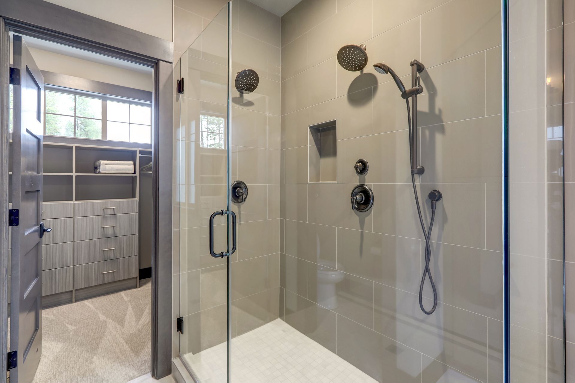Modern bathroom with glass shower, handheld showerhead, and tiled shower bench; vanity visible beyond.