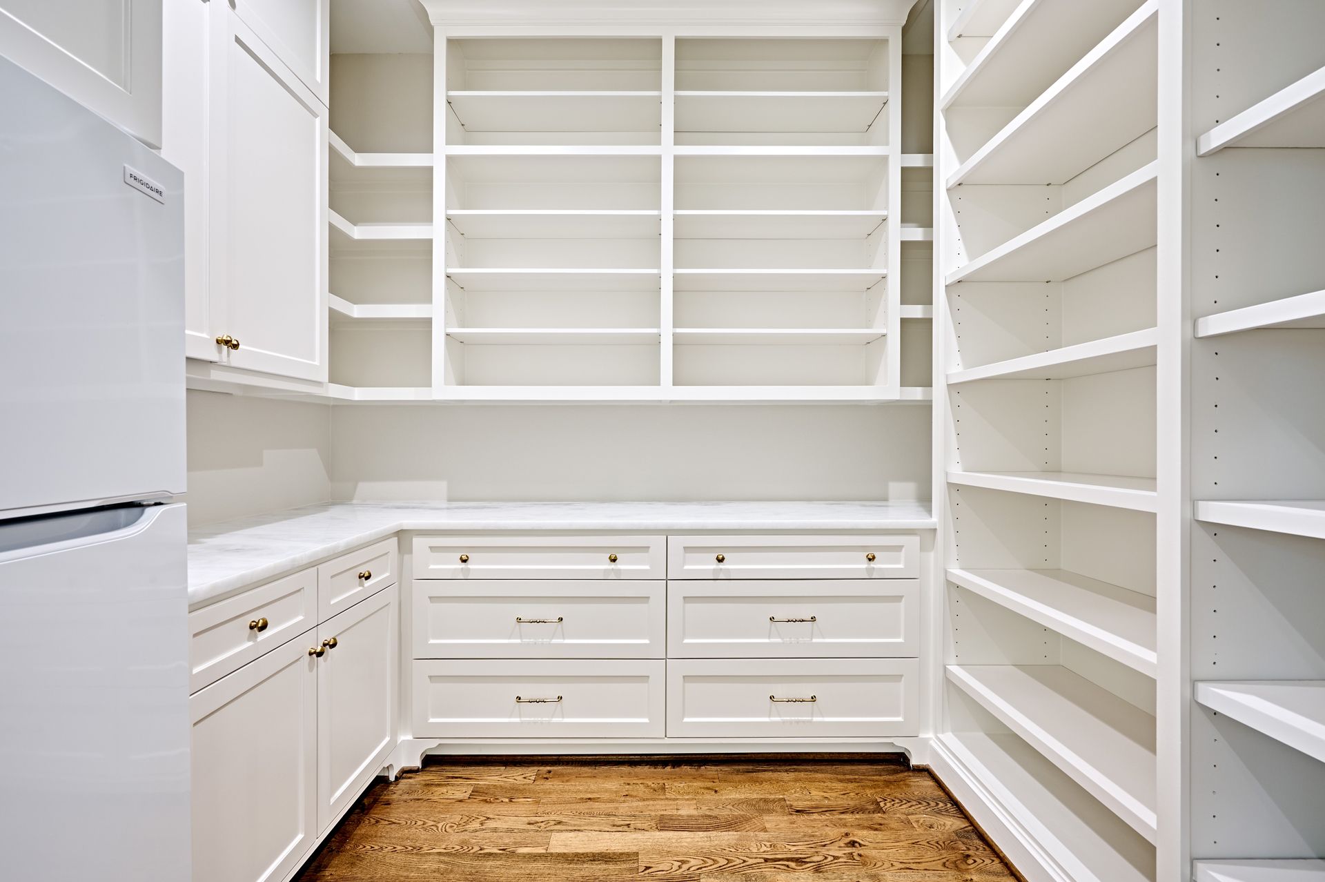 Bright white walk-in pantry with shelves, cabinets, drawers, and a wood floor