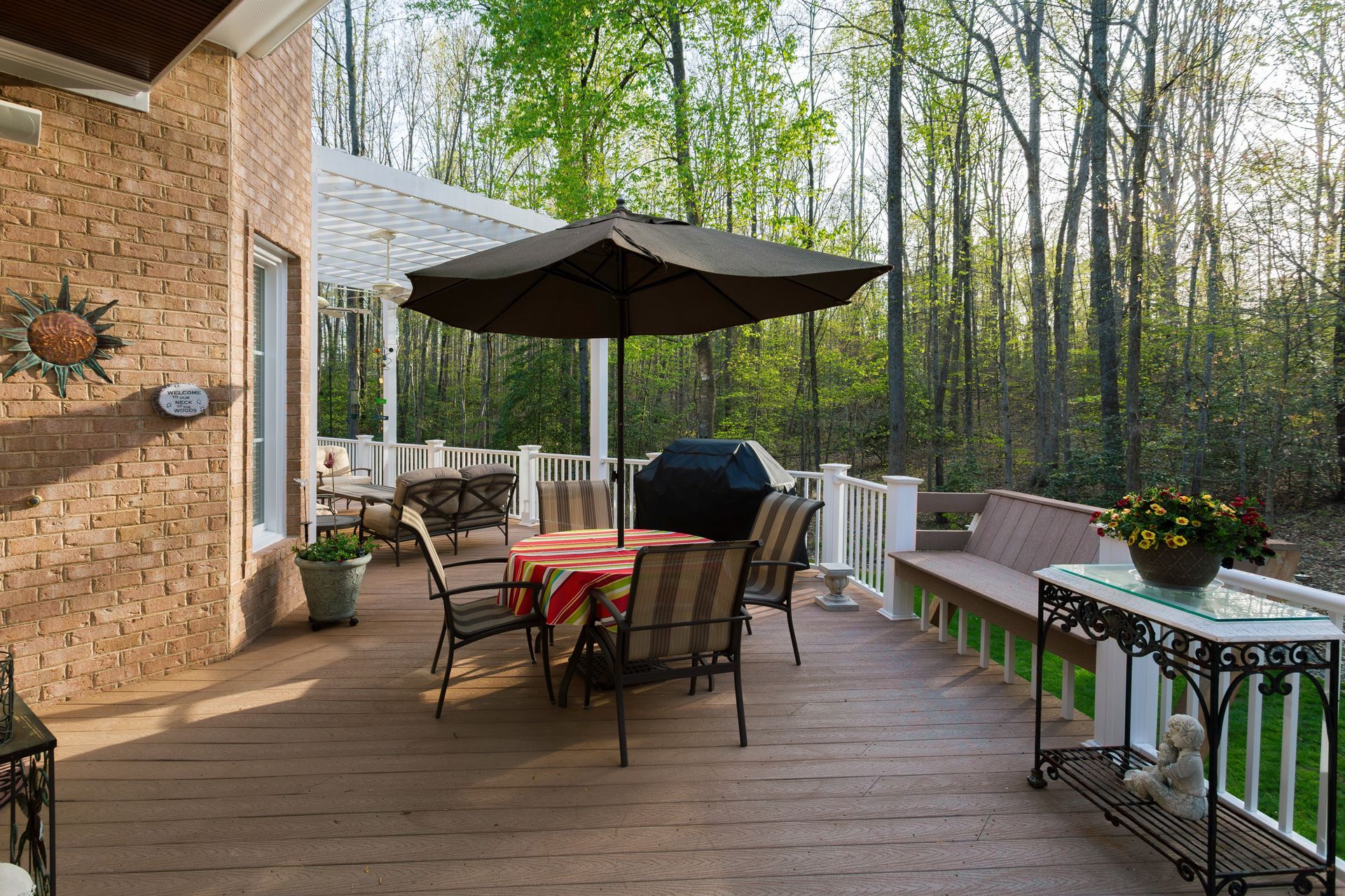 Backyard deck with table, chairs, umbrella, and potted flowers beside a brick house and wooded yard