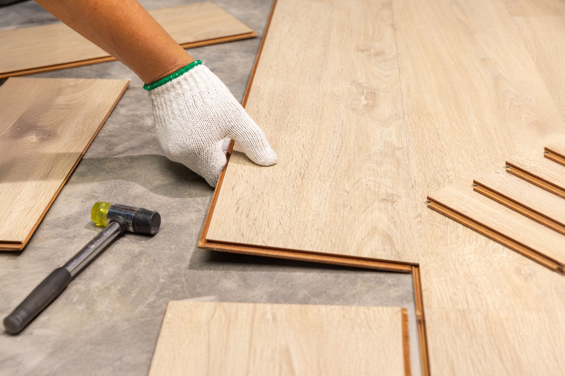 Gloved hand aligning wood floor planks beside a hammer and loose laminate pieces on a work surface