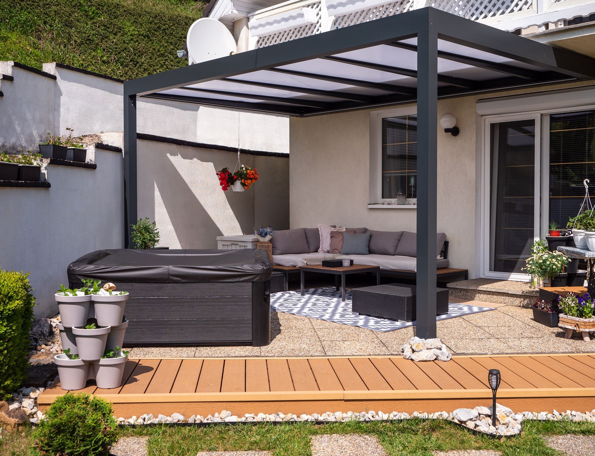 Modern patio with gray sectional sofa under a pergola, wooden deck, potted plants, and a dining area.