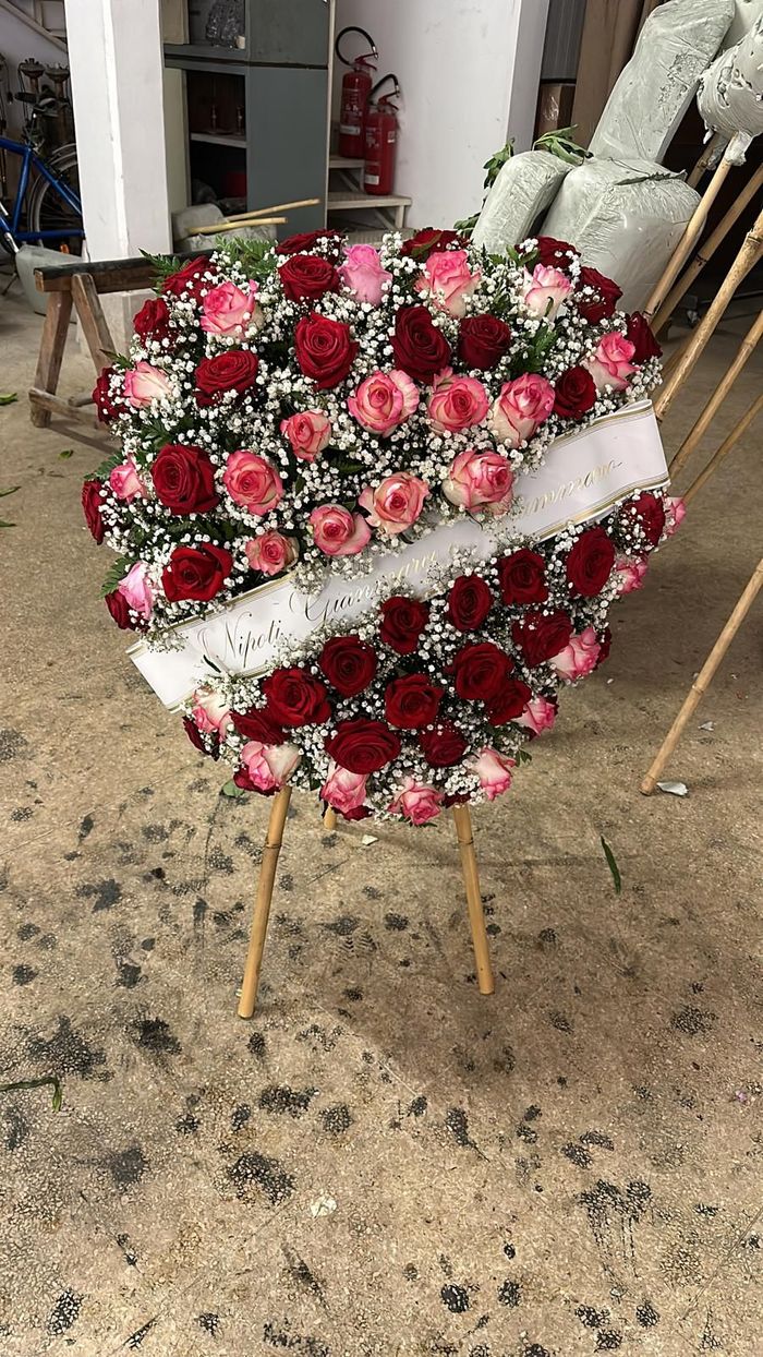 Composizione floreale a forma di cuore composta da rose rosse e rosa con nastro bianco, su un supporto.