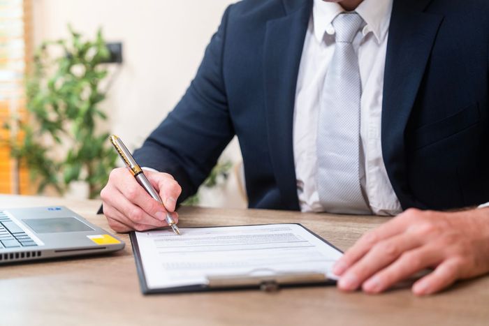 Uomo in giacca e cravatta che firma un documento su una lavagna, con il computer portatile sulla scrivania.