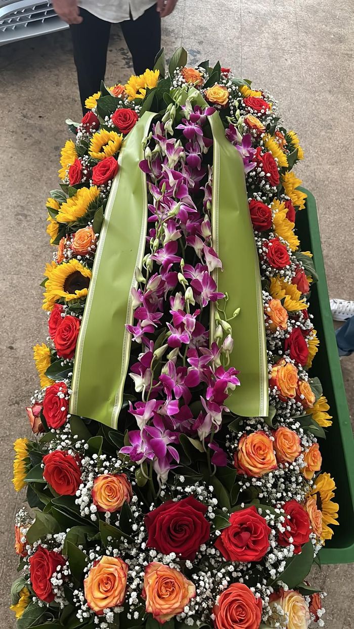 Composizione floreale su una bara, con girasoli, rose e orchidee con nastro verde.