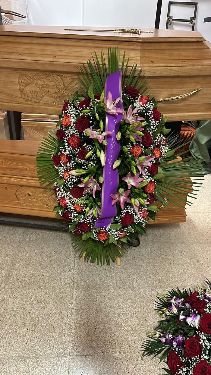 Composizione floreale con rose rosse, gigli e verde, su una bara di legno; nastro viola.