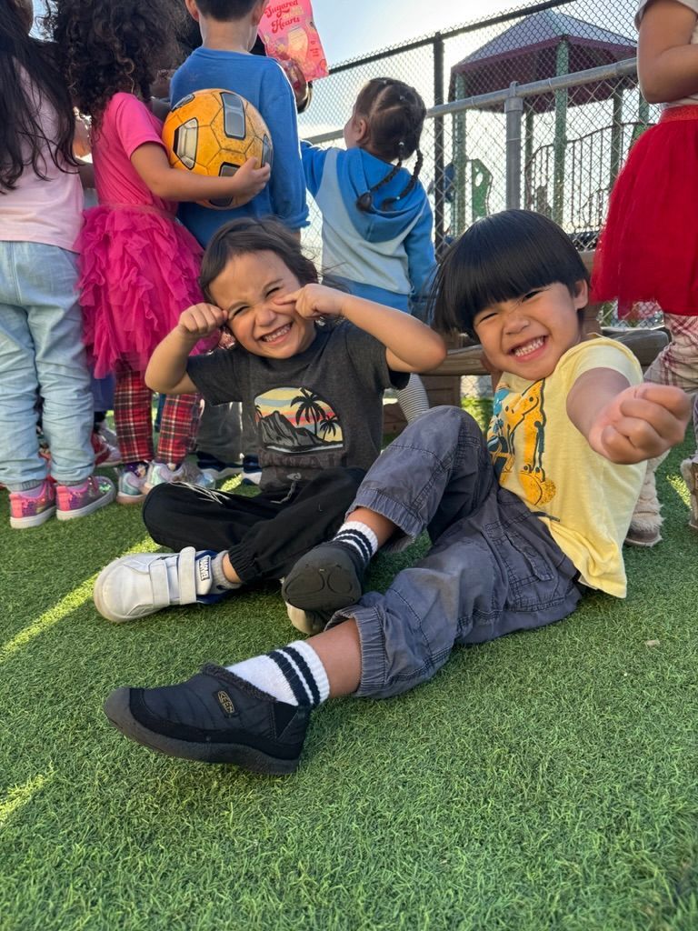 Two children drawing on paper attached to an easel in a classroom setting.