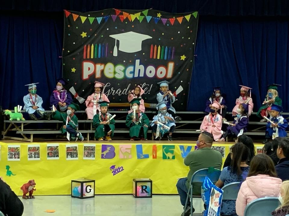 Preschool graduation ceremony with children in caps and gowns on stage. Parents watch from the audience.