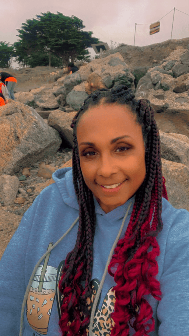 Woman smiles, wearing coral top and gray cardigan. Updo hairstyle, background of a stream.
